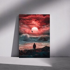 May include: A framed art print showing a dramatic seascape at sunset. The sky is a fiery red, with a large sun and silhouetted birds. A person stands on the rocky shore, facing the ocean with large waves.