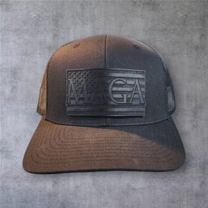 May include: A black trucker hat with a black leather patch featuring an American flag and the text "MAGA".