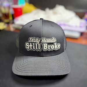 Może przedstawiać: Czarna czapka trucker z białym haftem z napisem "Dirty Hands Still Broke".