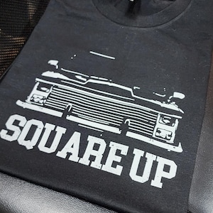Puede incluir: Camiseta negra con un gráfico blanco de un camión vintage y el texto "SQUARE UP".