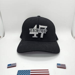 May include: A black baseball cap with a silver embroidered patch that says "TRUMP 47". There are two small American flags on the white surface below the cap.