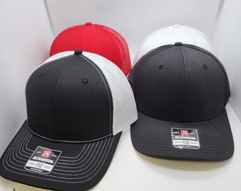May include: Three trucker hats in different colors: black with white mesh, red, and white. The hats have a black brim and a white stitching detail. The black hat has a white label with the text "Richardson 112 Original Trucker Adjustable".
