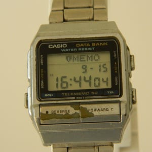 Może przedstawiać: Srebrny zegarek Casio Data Bank Telememo 50 z czarnym wyświetlaczem cyfrowym pokazującym godzinę 16:4404 i datę 9-15. Zegarek ma przyciski do regulacji, wyboru/trybu, sch, memo, ustawiania, światła i tel.