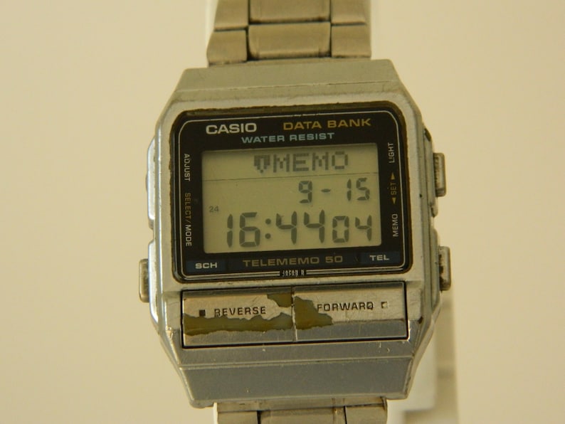 May include: A silver Casio Data Bank Telememo 50 wristwatch with a black digital display showing the time 16:4404 and the date 9-15. The watch has buttons for adjust, select/mode, sch, memo, set, light, and tel.