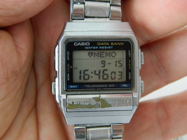 May include: A silver Casio Data Bank Telememo 50 digital watch with a black face and a silver metal band. The watch displays the time, date, and a memo. The watch has a variety of buttons, including adjust, select/mode, sch, tel, memo set, light, and forward.