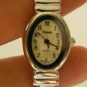 May include: Silver-toned quartz watch with an oval face. The watch face is off-white with black numerals and hands. The brand name "Xanadu" is printed above the center. The watch band is silver and has a flexible design.
