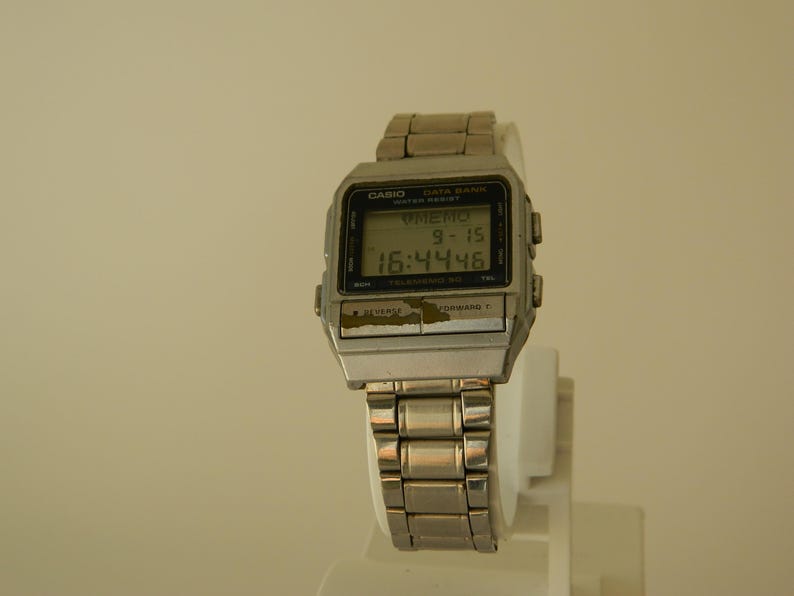 May include: A silver Casio Data Bank watch with a black digital display showing the time 16:44:46 and the date 9-15. The watch has a silver metal band.