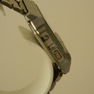 May include: A silver-toned wristwatch with a square face and a metal band. The watch has a digital display and a few buttons on the side.