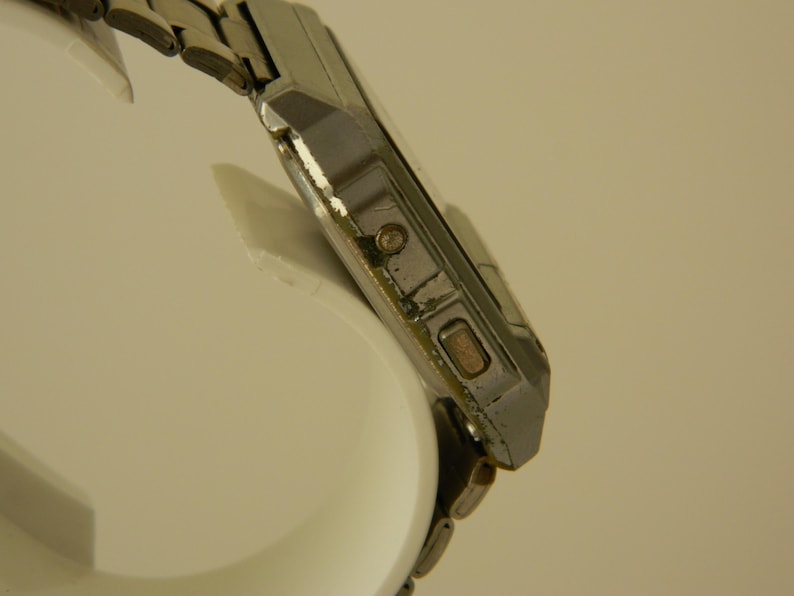 May include: A silver-toned wristwatch with a square face and a metal band. The watch has a digital display and a few buttons on the side.