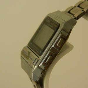 May include: A silver Casio digital watch with a rectangular face and a metal band. The watch has a digital display and several buttons on the side.