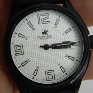 May include: A black wristwatch with a white face and black hands. The watch face has a textured pattern and the text "Beverly Hills Polo Club".