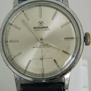 Könnte beinhalten: Eine silberne Rodania Armbanduhr mit weißem Zifferblatt und schwarzen Zeigern. Das Zifferblatt der Uhr hat schwarze Markierungen für Stunden und Minuten, und der Text "Rodania Uhrmachtswerk 17 Jewels Waterresistant" ist auf dem Zifferblatt aufgedruckt.