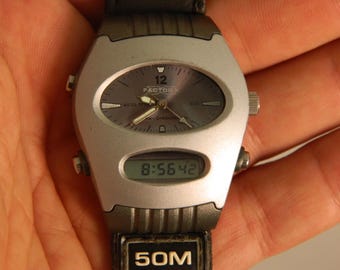 May include: A silver and black wristwatch with a digital display showing 8:56:42. The watch has a round analog face with a black background and white hands. The watch face also has the text "Factor" and "1/100 Chronograph" printed on it. The watch band is black with the text "50M" printed on it.