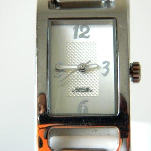 May include: Silver-toned wristwatch with a rectangular face. The watch face is silver with a textured center and displays the numbers 12, 3, and 6. The brand name "Dice Kayek" is visible. The watch has a silver band and a crown on the side.