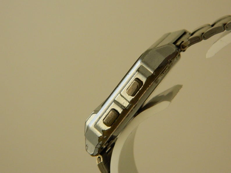 May include: A silver-toned wristwatch with a square face and a metal band. The watch has two buttons on the side of the case.