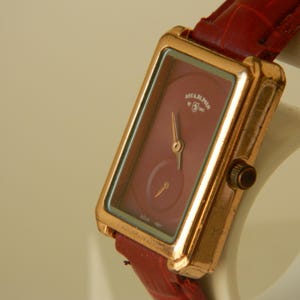 May include: A rectangular wristwatch with a gold-colored case and a burgundy face. The watch has a small seconds dial and is attached to a red leather strap. The watch face has the text "ARABELLE" and "SINCE 1987".