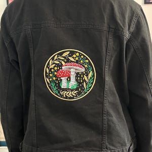 Può includere: Una giacca di jeans nera con una toppa ricamata di due funghi rossi e bianchi con foglie verdi e fiori gialli in un cerchio sul retro.