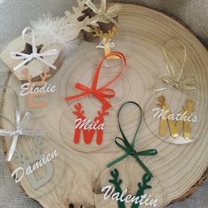 Könnte beinhalten: Klare Weihnachtsornamente aus Acryl mit Namen wie "Elodie", "Mila" und "Mathis" werden gezeigt. Jedes Ornament hat ein Rentier-Design und ist mit einem Band verziert. Die Ornamente sind auf einer Holzscheibe angeordnet und bilden eine festliche Präsentation.