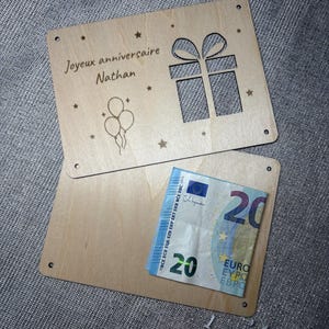 Könnte beinhalten: Ein hellbrauner Holz-Geschenkkartenhalter mit dem Text "Joyeux anniversaire Nathan" und einem ausgeschnittenen Geschenkbox-Design. Er enthält einen 20-Euro-Schein. Sterne und Ballons ergänzen das Design.