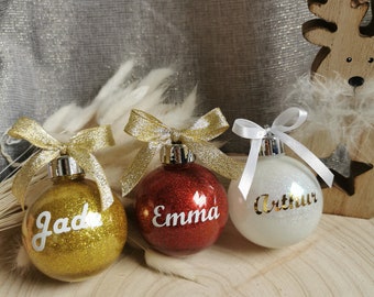Könnte beinhalten: Drei personalisierte Weihnachtskugeln. Eine ist goldfarben mit dem Namen "Jade", eine ist rot mit "Emma" und eine ist weiß mit "Arthur". Jede Kugel hat ein glitzerndes Finish und ist mit einer Schleife versehen. Die Kugeln liegen auf einer Holzoberfläche.