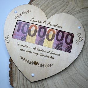 Könnte beinhalten: Eine herzförmige Plakette aus hellem Holz mit den eingravierten Namen Laura & Aurélien, der Zahl 1000000 und dem französischen Text Un million... de bonheur et d'amour pour votre magnifique union, mit einem Ausschnitt für violette Geldscheine.