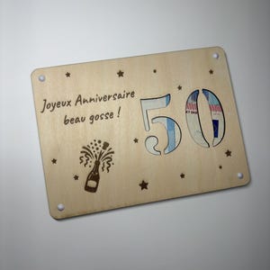 Könnte beinhalten: Eine rechteckige, helle Holzplatte mit dem lasergravierten Text Joyeux Anniversaire beau gosse! und einer Champagnerflaschen-Illustration. Die Zahl 50 ist ausgeschnitten und gibt den Blick auf dahinterliegende Geldscheine frei, verziert mit Sternen.