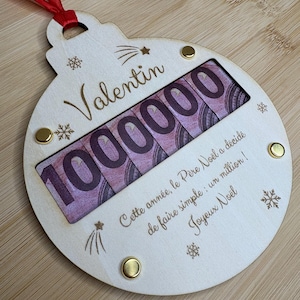 Peut inclure: Une décoration de Noël ronde en bois avec un ruban rouge, portant le prénom Valentin, le nombre 1000000 et le texte : Cette année, le Père Noël a décidé de faire simple : un million ! Joyeux Noël, agrémentée de gravures de flocons.
