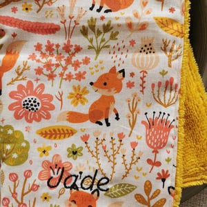 Peut inclure: Une couverture de bébé blanche avec une bordure jaune et un motif de renard et de fleurs. La couverture est personnalisée avec le nom "Jade" brodé en fil noir.