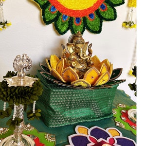 May include: A golden statue of Ganesha sits atop a lotus flower-shaped base, inside a green patterned container. A colorful peacock feather design hangs on the wall behind the statue. A silver lamp and decorative floral garlands are also visible.