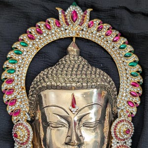 May include: A golden Buddha statue with intricate details. The statue features a serene face with closed eyes and a decorative halo adorned with red, green, and clear gemstones. The halo and earrings are embellished with sparkling stones.