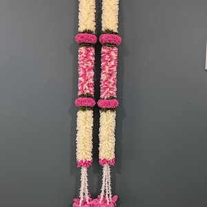 May include: Two vertical floral garlands with alternating sections of white and pink flowers. The garlands are adorned with vibrant pink flower accents and a cluster of pink flowers at the bottom. The background is a solid gray.
