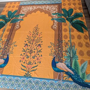 Puede incluir: Una alfombra de oraci&oacute;n rectangular con un fondo amarillo mostaza vibrante y un dise&ntilde;o de arco central. La alfombra muestra dos pavos reales azules entre palmeras verdes y un motivo floral central con detalles rosas y verdes.