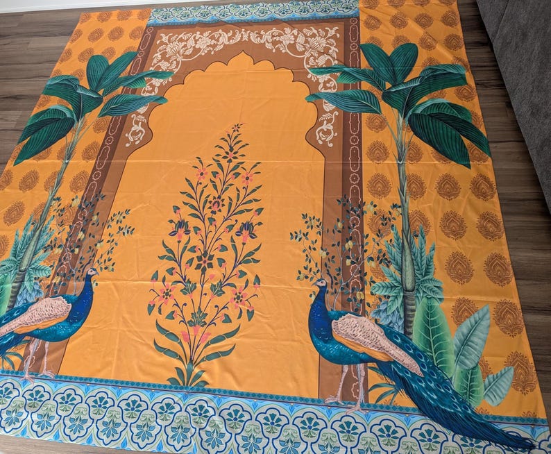 Puede incluir: Una alfombra de oraci&oacute;n rectangular con un fondo amarillo mostaza vibrante y un dise&ntilde;o de arco central. La alfombra muestra dos pavos reales azules entre palmeras verdes y un motivo floral central con detalles rosas y verdes.