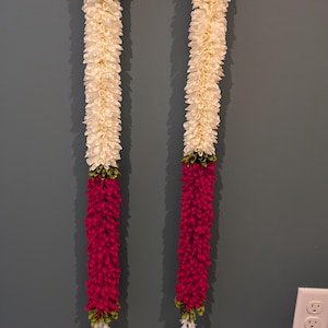 May include: Two decorative floral garlands. Each garland features alternating sections of cream-colored and deep red flowers. Green and white accents are visible at the top and bottom of each garland. The garlands are hanging against a gray wall.
