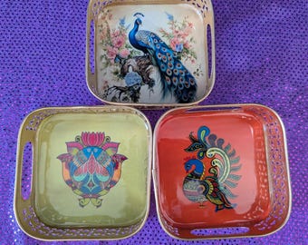 May include: Three decorative trays with gold filigree edges. One tray features a peacock with blue and green feathers, perched on a branch with pink flowers. Another has a colorful floral design, and the third has a peacock design.