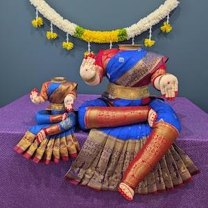 May include: Two traditional Indian dance costumes, one large and one small, in vibrant blue, red, and gold. The costumes are adorned with intricate patterns and embellishments, and are displayed on a purple surface.