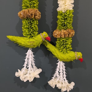 May include: Two decorative garlands with green and white floral elements. Each garland features a parrot made of green leaves, a red beak, and a jeweled collar. The garlands are adorned with gold accents and white floral pendants.