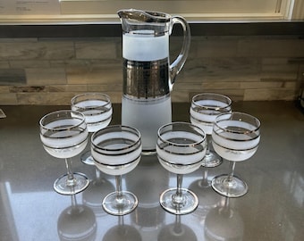 May include: A vintage glassware set featuring a clear glass pitcher with silver bands and a frosted base, accompanied by six matching wine glasses. The glasses and pitcher have a silver band design.