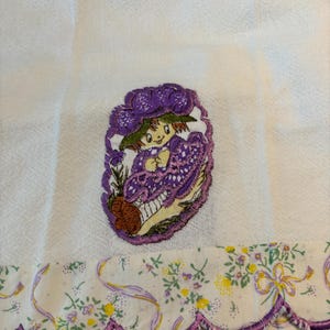 May include: A white tea towel with a purple embroidered design of a girl wearing a purple dress and hat. The towel has a scalloped edge with a floral print.