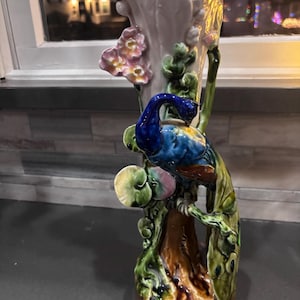 May include: A tall, ceramic vase with a peacock design. The vase features a light purple body with a turquoise interior and is adorned with a colorful peacock, pink flowers, and green foliage. The base is brown.