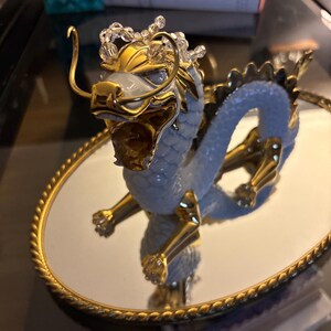 May include: A detailed figurine of a dragon with a gold-colored head, legs, and accents. The dragon has a light blue body and is posed on a round, mirrored tray with a gold-colored, rope-like border. The dragon's head is adorned with clear crystals.
