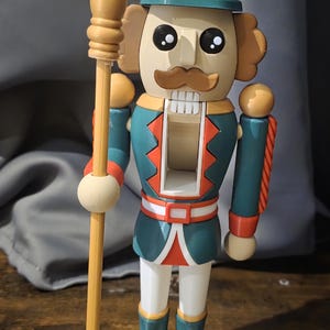 May include: A teal and red Christmas nutcracker figurine holding a wooden staff. The nutcracker has a candy cane on its hat and a mustache.