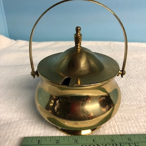 May include: A small, gold-colored brass sugar bowl with a lid and a handle. The bowl is about 4 inches tall and has a round, bulbous shape. The lid has a small knob on top and a small spout on the side. The handle is a simple loop that is attached to the sides of the bowl.