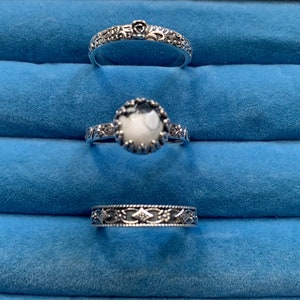 Può includere: Tre anelli d'argento con disegni intricati. L'anello centrale presenta una pietra bianca con un bordo argentato.