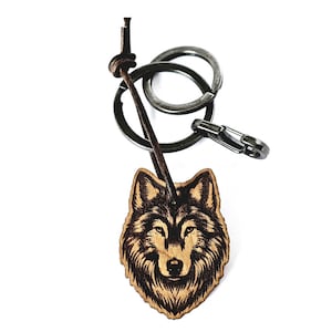 Könnte beinhalten: Ein Schlüsselanhänger aus Holz in Form eines Wolfskopfes mit einem schwarzen und braunen Design. Der Schlüsselanhänger ist an einem silbernen Schlüsselring aus Metall mit einer Lederschnur befestigt.