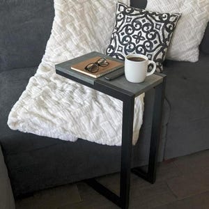 May include: A small, gray and black side table with a cup of coffee, a notebook, and glasses. The table is next to a gray couch with a white blanket and decorative pillow.