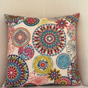 Puede incluir: Una almohada blanca con un patrón de mandala colorido. El patrón presenta colores rojo, azul, verde y rosa. La almohada está hecha de una tela suave y cómoda.