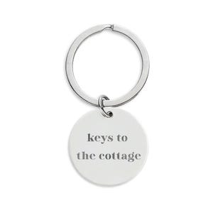 Op de afbeelding: Een zilverkleurige sleutelhanger met een ronde, witte hanger. De hanger heeft de tekst "keys to the cottage" in een grijze, schreefloze lettertype. De sleutelring is cirkelvormig en met een kleine lus aan de hanger bevestigd.