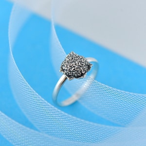 May include: A silver ring with a textured, heart-shaped design. The ring is set against a light blue background with a white ribbon. The ring's band is thin and delicate.