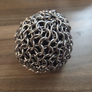 Puede incluir: Objeto esférico cubierto con un patrón similar a una cota de malla. Los eslabones metálicos plateados crean una superficie texturizada. El objeto está sobre una superficie de madera. La forma es redonda y compacta.